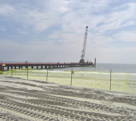 A sandy beach with tire tracks leading to a long pier extending into the ocean. A crane is positioned on the pier. The sky is partly cloudy, and the water appears calm. A fence runs along the beach, parallel to the shoreline.