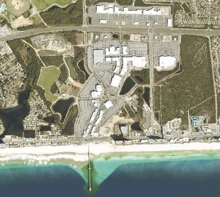 Aerial view of a coastal town showing a beachfront with a pier extending into the ocean. There are buildings, parking lots, and green spaces behind the beach. Roads and residential areas are visible further inland.