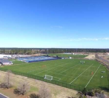 Aerial view of a sports complex with green soccer fields and fenced tennis courts. A few soccer goals are scattered on the fields. The area is surrounded by trees and bordered by a road. The sky is clear and blue.