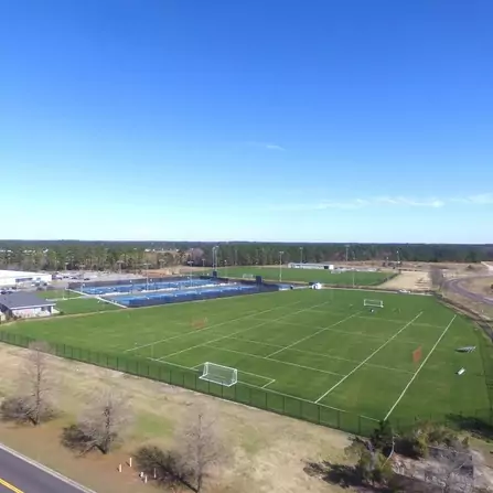 Aerial view of a sports complex with green soccer fields and fenced tennis courts. A few soccer goals are scattered on the fields. The area is surrounded by trees and bordered by a road. The sky is clear and blue.