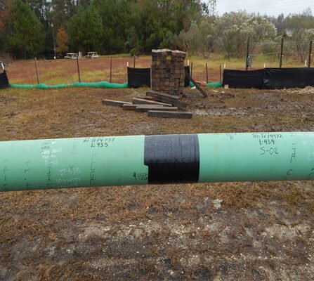 A large green pipeline section with markings and a black patch is placed on the ground at a construction site. Behind it, there are stacks of bricks, wooden planks, and green grass bordered by a black mesh fence and trees.