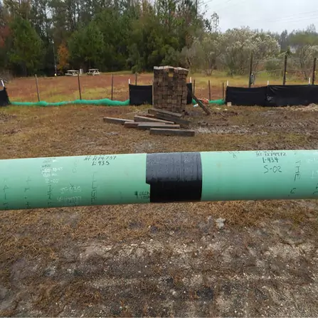 A large green pipeline section with markings and a black patch is placed on the ground at a construction site. Behind it, there are stacks of bricks, wooden planks, and green grass bordered by a black mesh fence and trees.