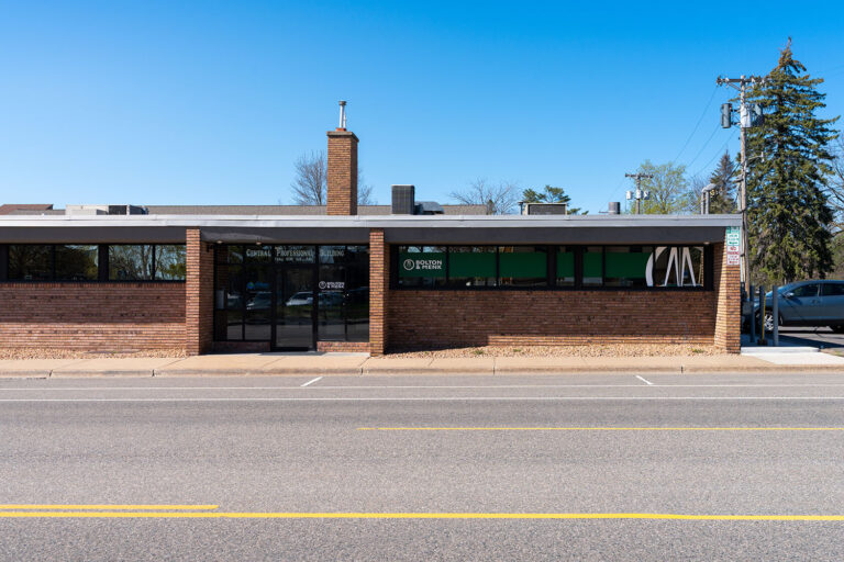 Single-story commercial building with a brick facade and large windows, labeled Dental Professional Building. A road and parking area are in the foreground, with trees and a neighboring building in the background under a clear sky.