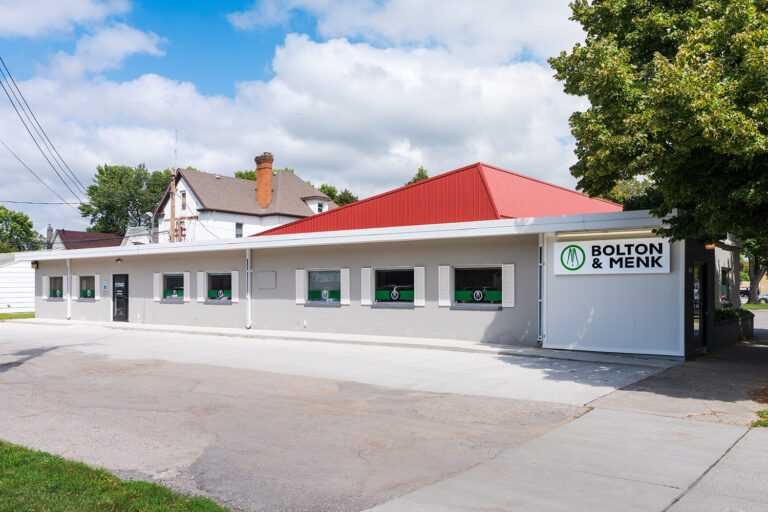A small, modern office building with a red roof and white-gray exterior features the Bolton & Menk signage prominently. This Bolton & Menk location is accented by several windows, basking in sunlight under a lightly clouded sky.