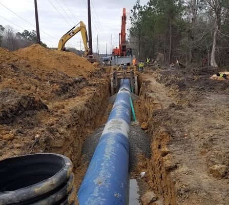 Construction site with workers installing a large blue pipe in a trench. Excavators are visible, and surrounding areas have piles of dirt and forested land. Power lines run parallel to the trench.