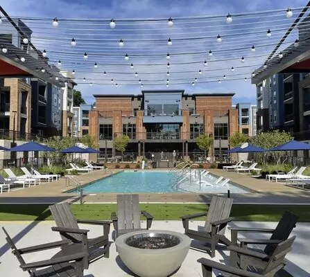 A modern outdoor pool area at an apartment complex. The scene includes a central pool, surrounded by lounge chairs with blue umbrellas, a round fire pit with wooden chairs, and string lights overhead. A multi-story building is in the background.