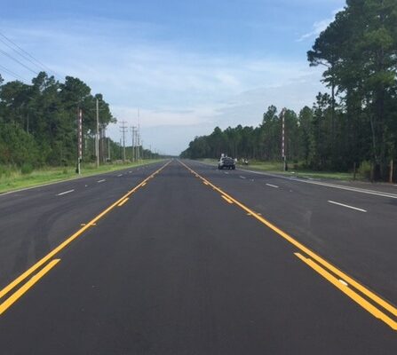 A wide, freshly paved road stretches into the distance, bordered by dense trees on either side. Clear blue sky above with yellow lane markings on the road. A car is visible in the distance driving along the road.