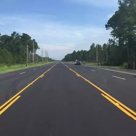 A wide, freshly paved road stretches into the distance, bordered by dense trees on either side. Clear blue sky above with yellow lane markings on the road. A car is visible in the distance driving along the road.