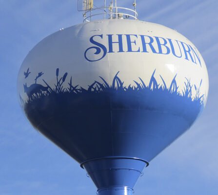 A water tower painted white and blue with SHERBURN written at the top. The lower half features silhouettes of grass, cattails, and animals like a deer and a bird against a blue backdrop under a clear sky.