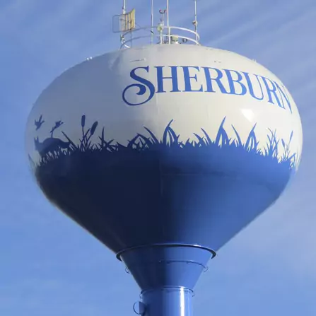 A water tower painted white and blue with SHERBURN written at the top. The lower half features silhouettes of grass, cattails, and animals like a deer and a bird against a blue backdrop under a clear sky.