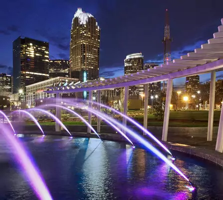 A nighttime cityscape featuring a series of fountains illuminated with purple lights, spraying water in arcs over a pool. Skyscrapers with lit windows and a distinct white structure in the foreground enhance the urban scene.
