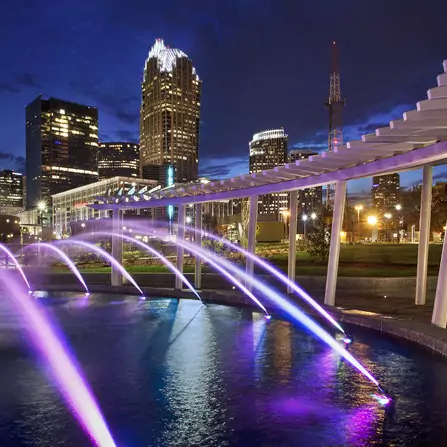 A nighttime cityscape featuring a series of fountains illuminated with purple lights, spraying water in arcs over a pool. Skyscrapers with lit windows and a distinct white structure in the foreground enhance the urban scene.