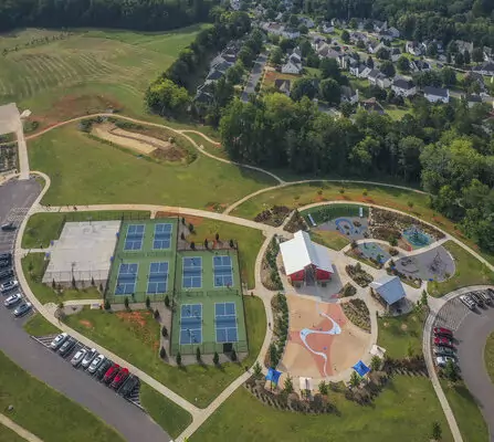 Aerial view of a recreational park with tennis courts, a playground, and a splash pad. A parking lot is on the left, and a residential neighborhood and forested area are in the background. Pathways connect the different areas of the park.