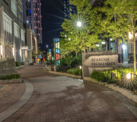 Night view of Bearden Promenade, featuring a well-lit brick pathway lined with trees and modern buildings. Several colorful glass installations and bright streetlights create a vibrant atmosphere.