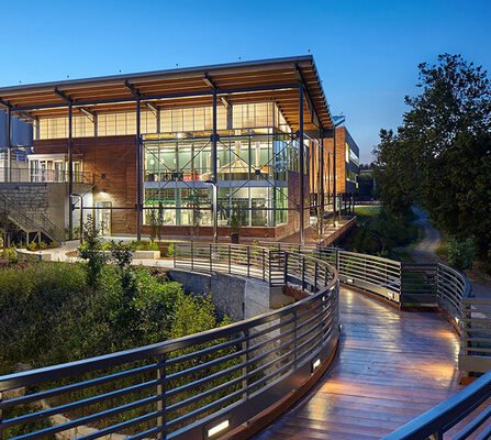 A modern building with large glass windows and wooden accents is illuminated at dusk. A curved walkway with metal railings leads to the entrance, surrounded by greenery and trees under a clear blue sky.