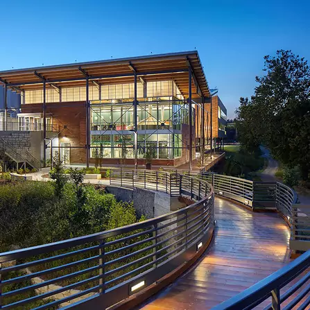 A modern building with large glass windows and wooden accents is illuminated at dusk. A curved walkway with metal railings leads to the entrance, surrounded by greenery and trees under a clear blue sky.