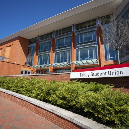 Exterior view of a modern building with large glass windows and red brick walls. A sign reads Talley Student Union. The foreground features a low hedge and a brick pathway, under a clear blue sky.