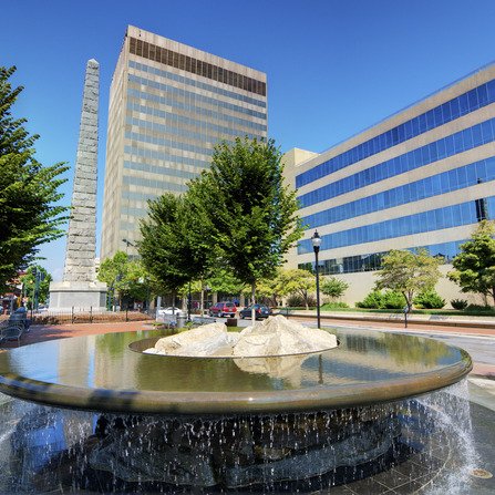 A circular fountain with cascading water and large rocks in the center. Behind it, a tall obelisk stands next to modern office buildings. Lush trees line the pathway under a clear blue sky.