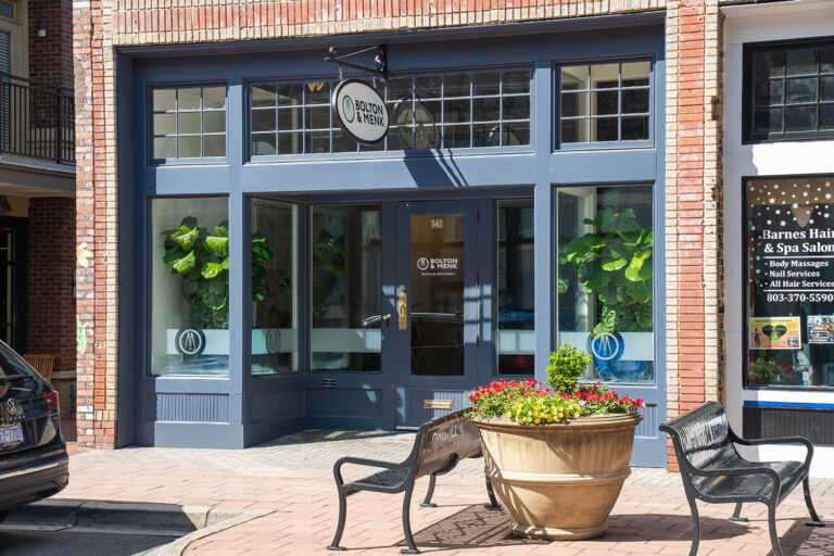 A brick building with large, blue-framed windows houses a tranquil spa salon, reminiscent of Bolton & Menk locations. A car is parked nearby, while two benches sit beside a flower-filled planter. The salons sign highlights its luxurious hair and spa services.
