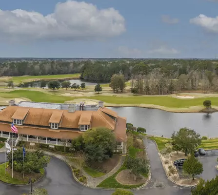 Aerial view of a golf course with a clubhouse in the foreground. The clubhouse has a rust-colored roof and an American flag nearby. The course features lush green fairways, sand traps, and a pond. Trees and a cloudy sky are in the background.