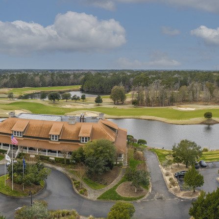 Aerial view of a golf course with a clubhouse in the foreground. The clubhouse has a rust-colored roof and an American flag nearby. The course features lush green fairways, sand traps, and a pond. Trees and a cloudy sky are in the background.