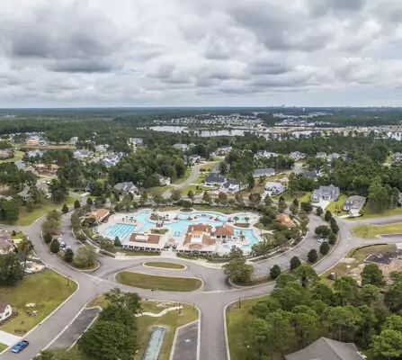 Aerial view of a suburban neighborhood featuring a circular layout with a large community pool and several surrounding buildings. Streets radiate outward, bordered by houses and trees. Cloudy sky overhead.