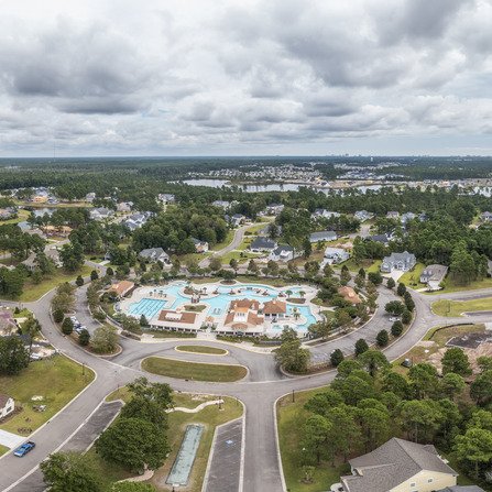 Aerial view of a suburban neighborhood featuring a circular layout with a large community pool and several surrounding buildings. Streets radiate outward, bordered by houses and trees. Cloudy sky overhead.