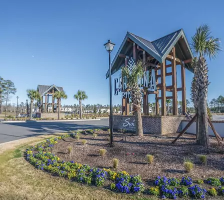 Entrance to a planned community with two modern wooden and brick structures featuring signage. Palm trees line the road, and colorful flowers are planted in the foreground. A clear blue sky is overhead.