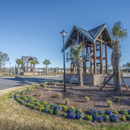 Entrance to a planned community with two modern wooden and brick structures featuring signage. Palm trees line the road, and colorful flowers are planted in the foreground. A clear blue sky is overhead.