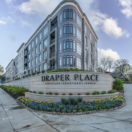 Four-story apartment building with large windows and Draper Place Signature Apartment Homes sign in front. Surrounded by landscaped gardens and blue sky with clouds above.
