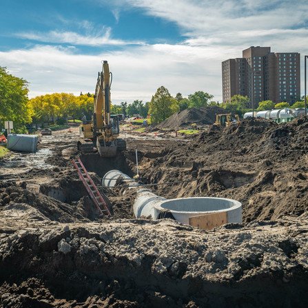 Water drainage area under construction with pipe and digger, tall building in the background.