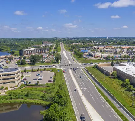 Aerial view of a highway cutting through a mix of residential areas, offices, large buildings, and a lush wooded area