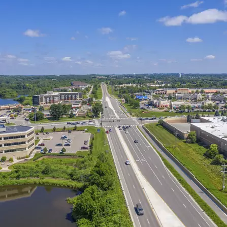 Aerial view of a highway cutting through a mix of residential areas, offices, large buildings, and a lush wooded area