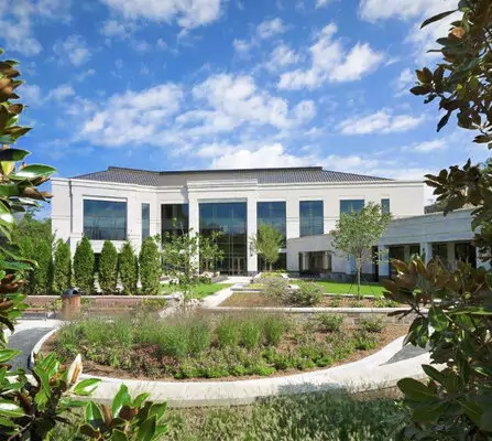 A modern two-story building with large windows, surrounded by landscaped gardens and framed by trees. The sky is partly cloudy, and a curved pathway leads to the entrance.