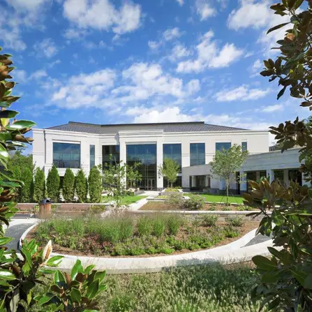 A modern two-story building with large windows, surrounded by landscaped gardens and framed by trees. The sky is partly cloudy, and a curved pathway leads to the entrance.