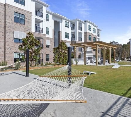 Contemporary apartment building with large windows and balconies. In the foreground, a hammock is stretched between poles on a patio, and theres a pergola with benches in a grassy area. The sky is clear and blue.