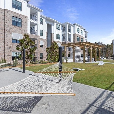 Contemporary apartment building with large windows and balconies. In the foreground, a hammock is stretched between poles on a patio, and theres a pergola with benches in a grassy area. The sky is clear and blue.