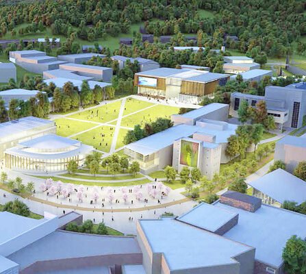 Aerial view of a university campus with modern buildings surrounding a large central green lawn. Pathways crisscross the lawn. Trees and hills are in the background, creating a serene and natural setting.