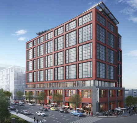 A modern multi-story building with red and black facade elements stands at a busy urban intersection, with cars and pedestrians passing by. The structure features large grid-patterned windows. The sky is clear with a few clouds.