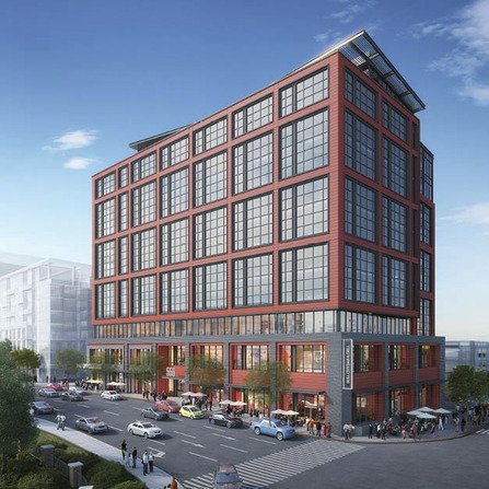 A modern multi-story building with red and black facade elements stands at a busy urban intersection, with cars and pedestrians passing by. The structure features large grid-patterned windows. The sky is clear with a few clouds.