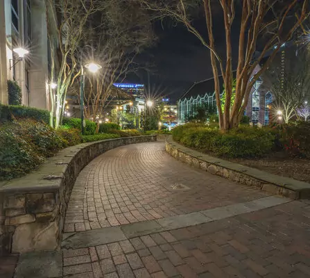 Brick walkway winding through a landscaped area at night, bordered by leafless trees and illuminated by streetlights. Buildings in the background have lights, enhancing the serene urban atmosphere.