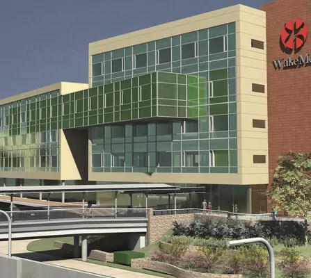 Modern building featuring large glass windows and green accents. The structure has multiple levels, with an overpass in the foreground. There is a logo on one side of the building, and landscaping with greenery around the entrance.