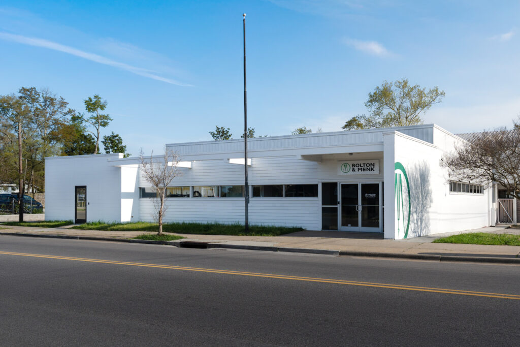 A modern white building with flat roofs and large windows, reminiscent of Bolton & Menk locations, stands amidst lush grass and a few trees. A blue mailbox marks the street corner under a clear sky dotted with a few clouds.