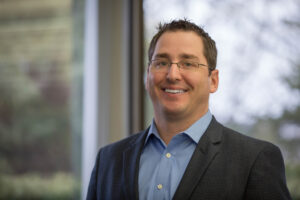 Smiling man with short dark hair and glasses wearing a blue shirt and dark blazer, standing by a large window. The blurred greenery outside adds to the serene ambiance as he reflects on how MATHCOUNTS creates positive experiences through challenging yet rewarding problem-solving activities.