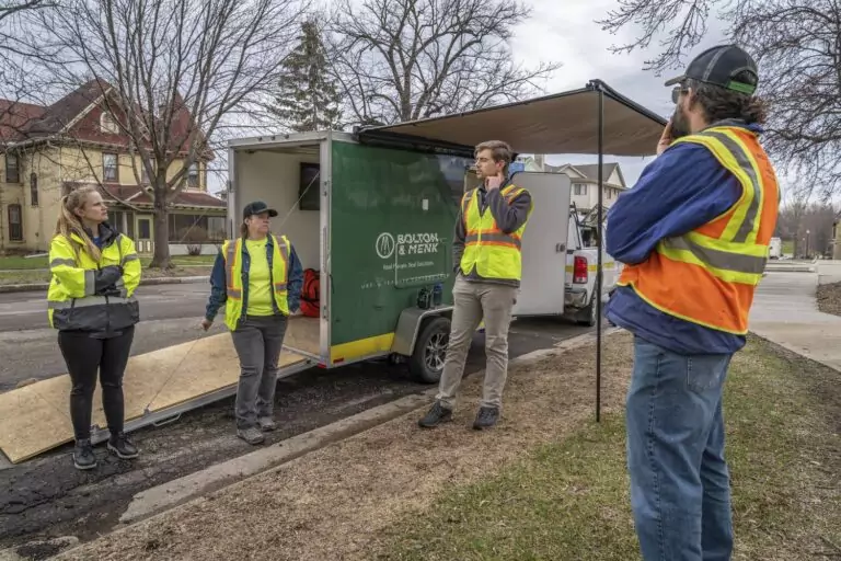 Four people in safety vests stand near a trailer labeled Bolton & Menk on a residential street. As part of local apprenticeship programs, three engage in conversation while one looks on. The open trailer door reveals equipment inside, with leafless trees and houses in the background.