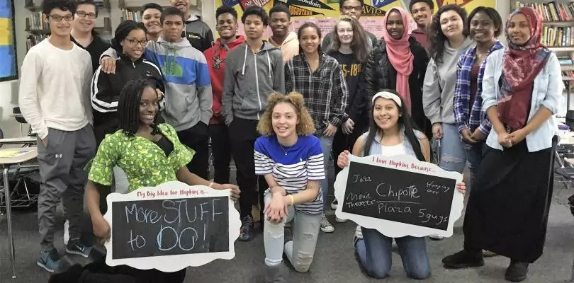 A diverse group of students poses in a classroom, embodying the spirit of innovation. Two students kneel in front, holding signs with handwritten messages about infrastructure solutions. Bookshelves and posters are visible in the background, creating a casual and friendly atmosphere.