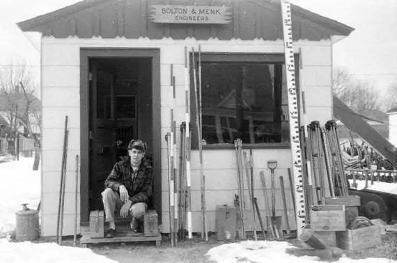 A person wearing a plaid shirt and cap sits on the snowy steps of Bolton & Menk Engineers, surrounded by measuring tools and equipment. The scene captures an expert poised to deliver innovative infrastructure solutions, with a large vertical measuring stick hinting at precision and expertise.