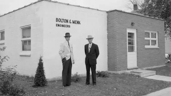 Two men in suits and hats stand before a small brick building labeled Bolton & Menk Engineers. Emphasizing their commitment to infrastructure solutions, the scene, set in a past era, includes a door, windows, and a nearby shrub.