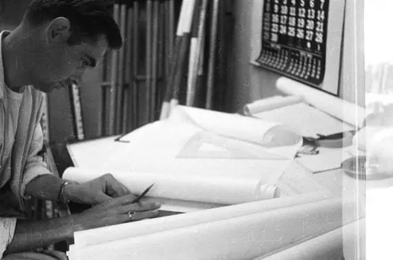 A man sits at a drafting table, intently sketching infrastructure solutions with a pencil. Rolls of paper and drafting tools are scattered around, evidence of his meticulous work. A calendar hangs on the wall in the background, completing the black and white scene.