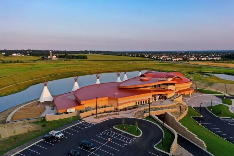 Aerial view of a large, circular building with a red roof, embodying traditional architecture. Its nestled amidst infrastructure solutions like a parking lot and fields, beside a serene pond. Teepee-like structures gracefully align at the back of the building.